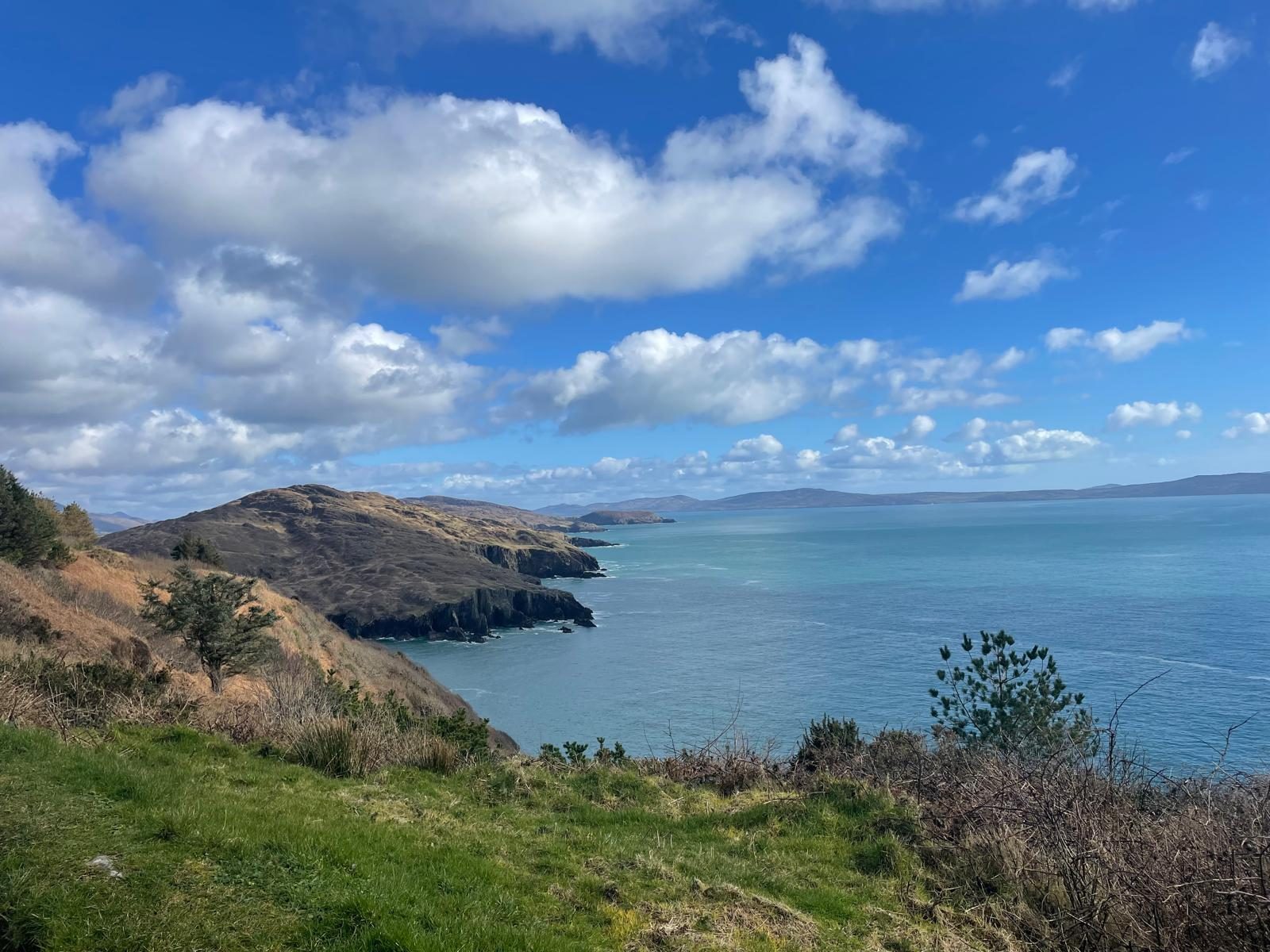 A green coastal headland meeting a calm turquoise sea under a sky of soft clouds