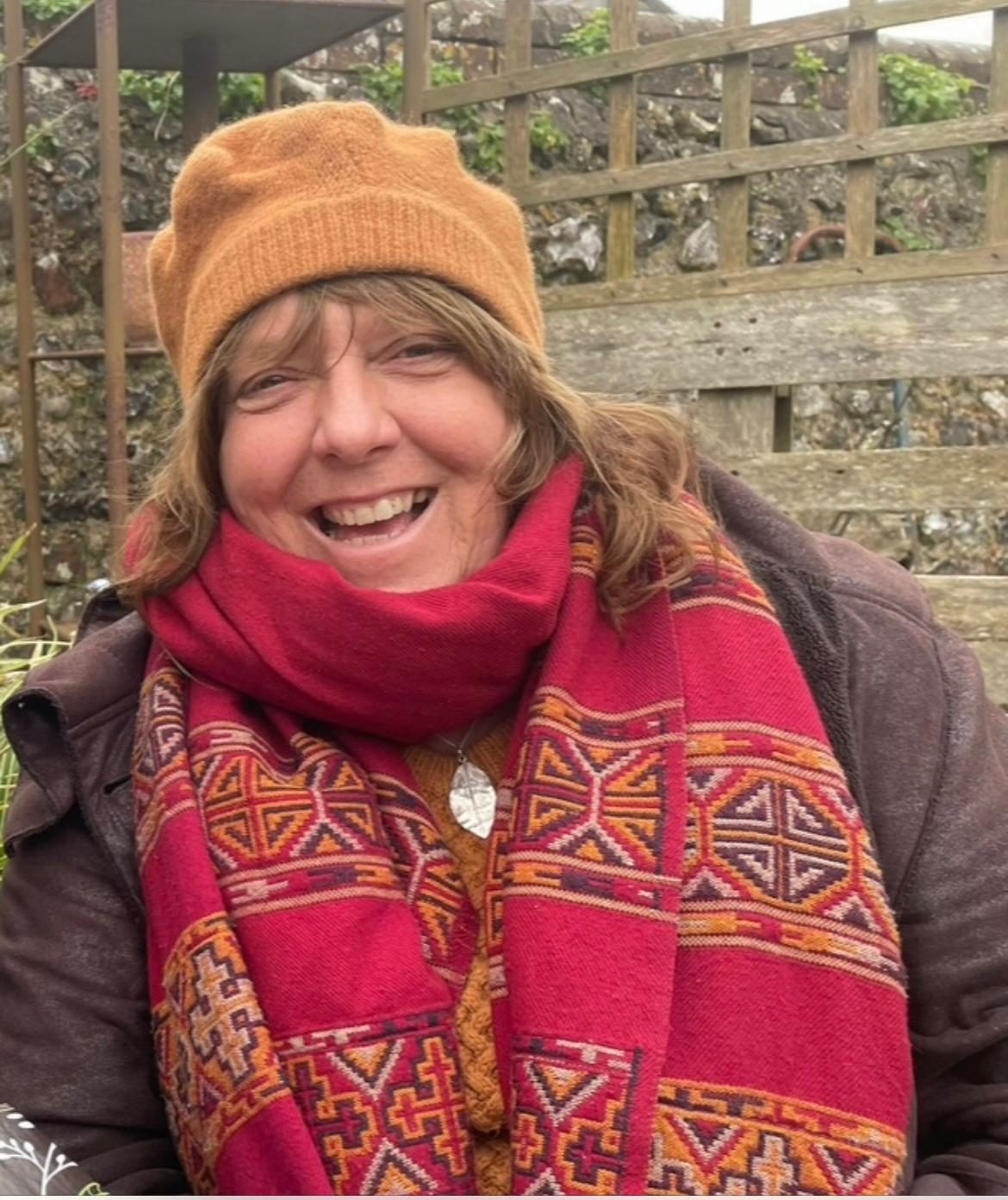 Lynne Christine Ridden smiling in her garden, wearing a warm hat and a red patterned scarf
